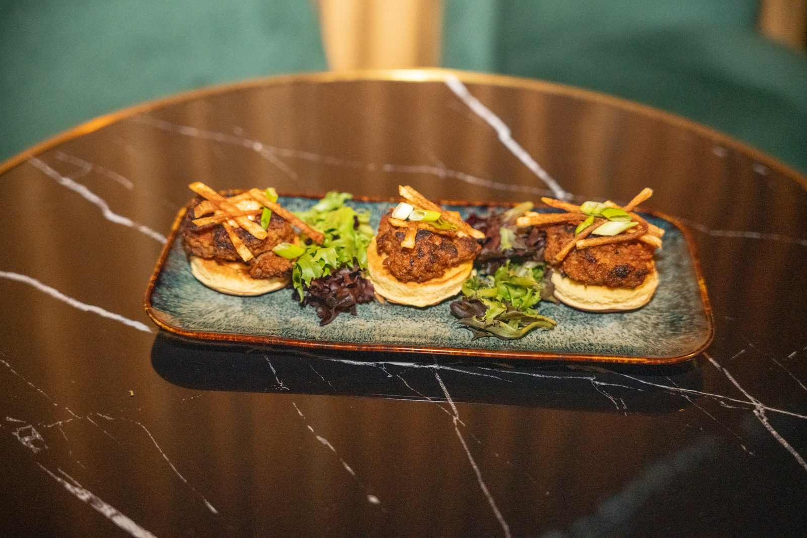 Three fried chicken biscuit sliders topped with crispy strips and scallions on a teal plate at Spanglish Martini Bar in Wynwood, Miami